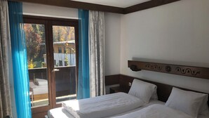 Habitación de hotel con dos camas, un cabecero de madera, vistas a un balcón con cortinas y una mesita de noche pequeña.
