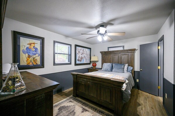 A bedroom with a wooden bed, a ceiling fan, framed artwork on the walls, and a nightstand with a lamp.