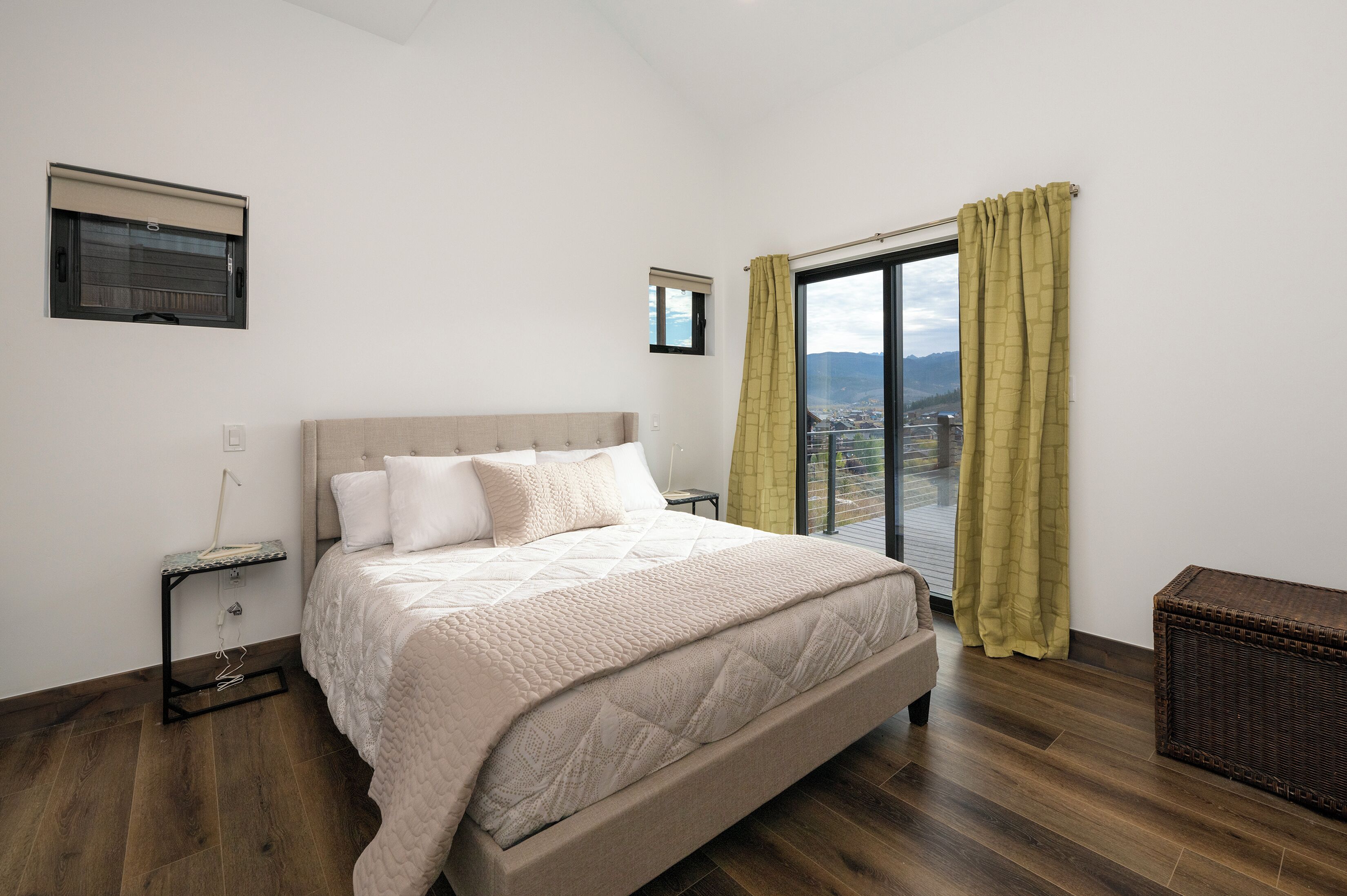 A bedroom with a large bed, a window with curtains, a small table, and a wicker chest.