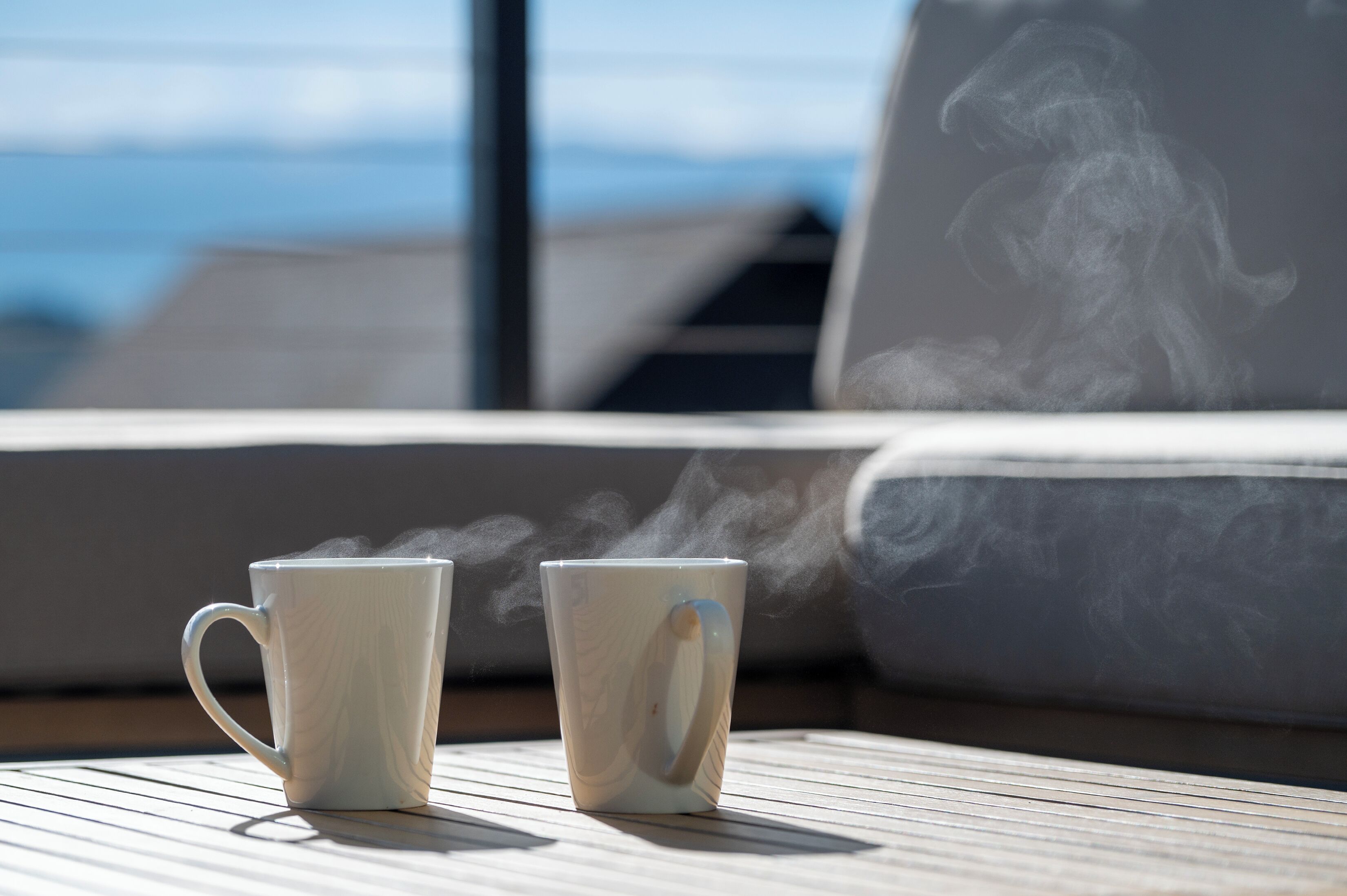 Zwei weiße Keramiktassen auf einem Holztisch, aus denen Dampf aufsteigt, auf einem Balkon mit Blick auf das Meer und die dahinterliegenden Berge.