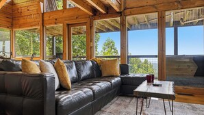 Ein Interieur einer Holzhütte mit einem Ledersofa, einem Tisch und großen Fenstern mit Blick auf Bäume.