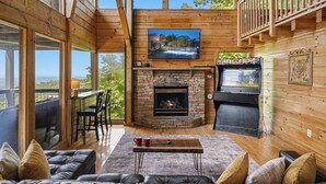 Ein gemütliches Cabin-Interieur mit Kamin aus Stein, einem darüber montierten Fernseher und einem Balkon mit Aussicht.