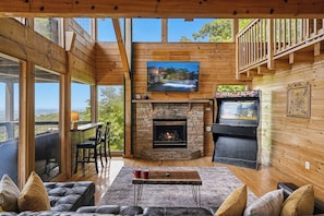 A cozy cabin interior with a stone fireplace, a television mounted above it, and a balcony with a view.