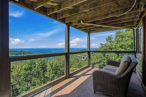 A covered balcony with a wicker chair, a wooden ceiling, and a view of a forested landscape.