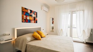 Ein Hotelzimmer mit einem großen Bett, einem Nachttisch, einer Wand-Klimaanlage, einem Fenster mit leichtem Vorhang und einem farbenfrohen abstrakten Gemälde.