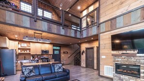 A spacious loft-style living area with a fireplace, a kitchen in the background, and a balcony with a view. - New Lux 4 King Suites | Hot Tub | Game Rm | PS5 (Gatlinburg)