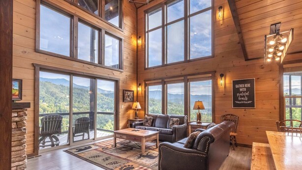 A spacious wooden interior with large windows offering mountain views, a cozy seating area, and a dining table. - New Lux 4 King Suites | Hot Tub | Game Rm | PS5 (Gatlinburg)