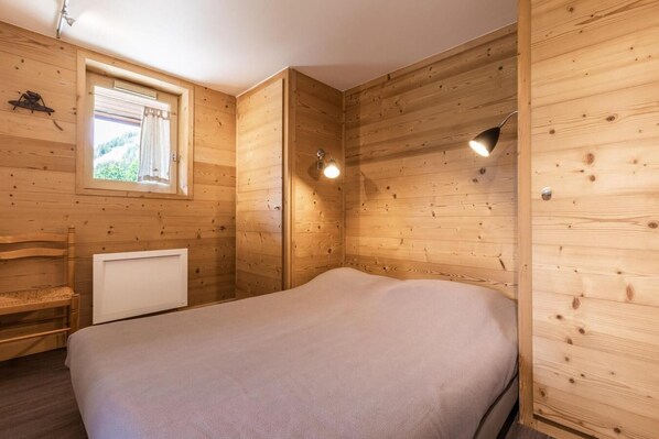 A wooden cabin room with a bed, a window with curtains, a chair, and a radiator.