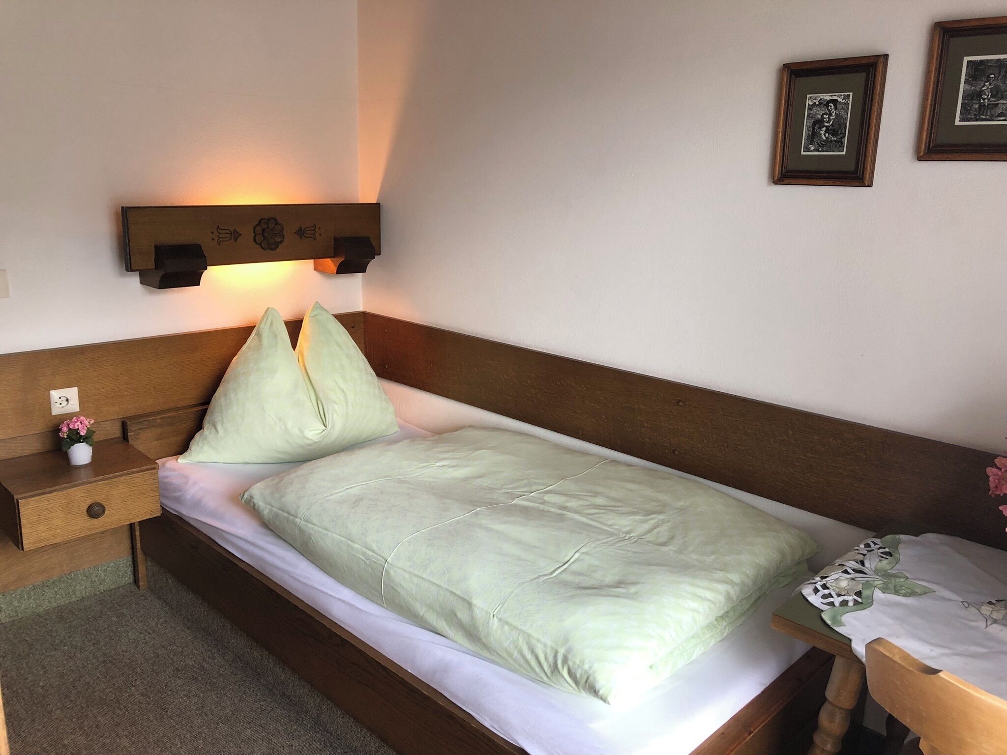 Ein Einzelbett mit Holz-Kopfstück, ein Nachttisch mit Lampe und ein kleiner Topfpflanze.