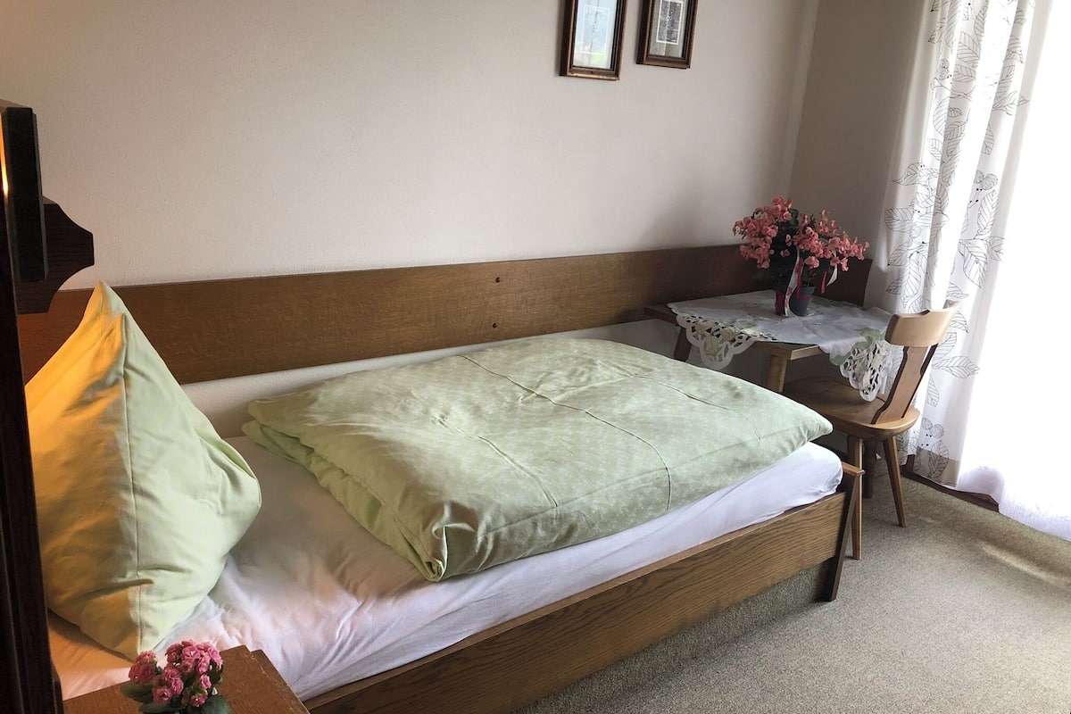 Una cama bien hecha con cabecera de madera, una mesita de noche con un jarrón de flores y una silla con cojín estampado.