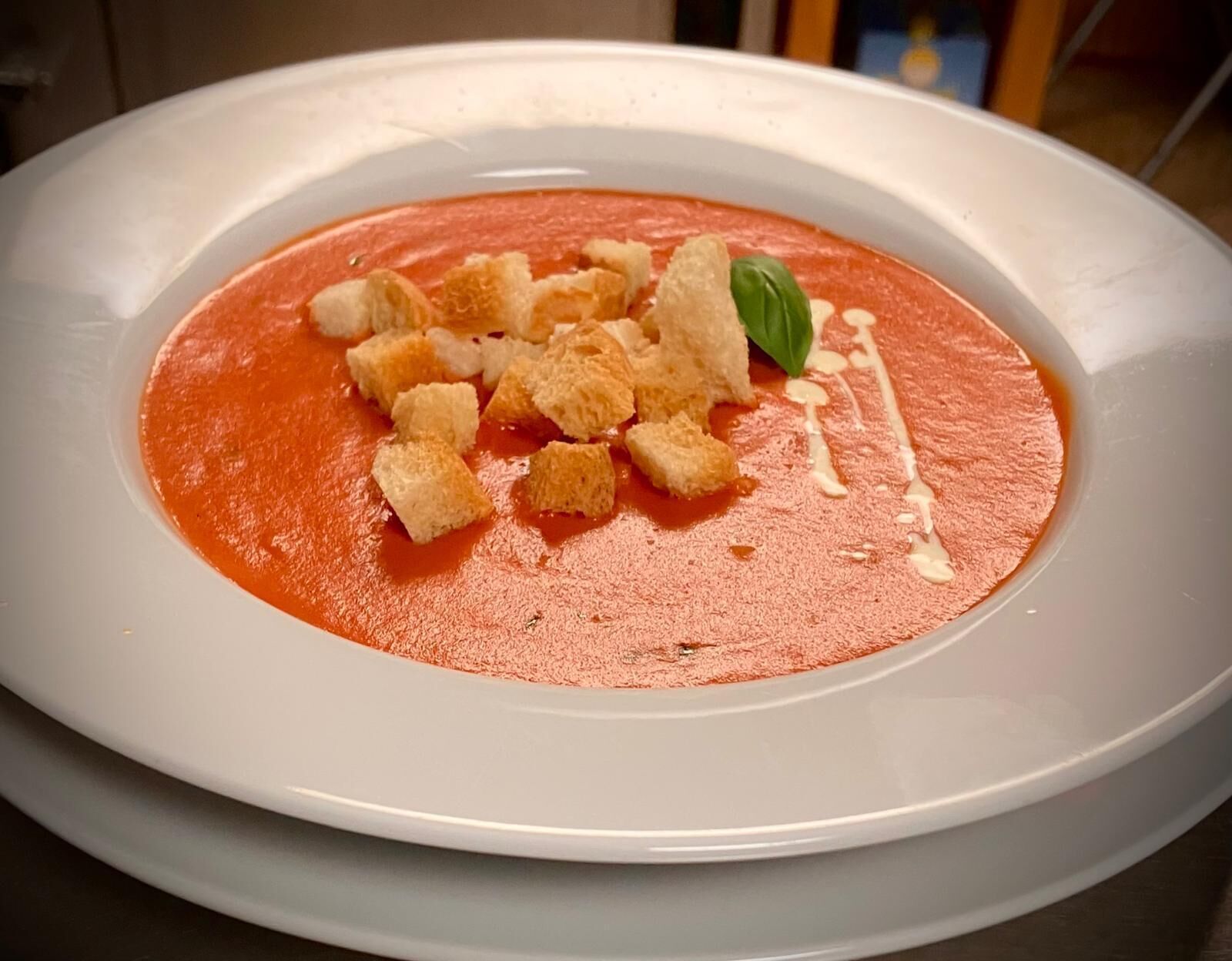 A white plate with a red soup, croutons, and a basil leaf garnish.