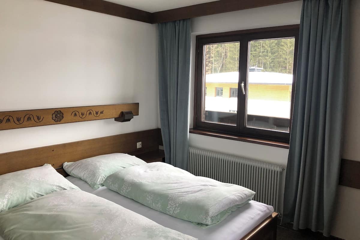 Una habitación con dos camas individuales, un cabecero de madera con detalles tallados, una ventana con cortinas y un radiador.