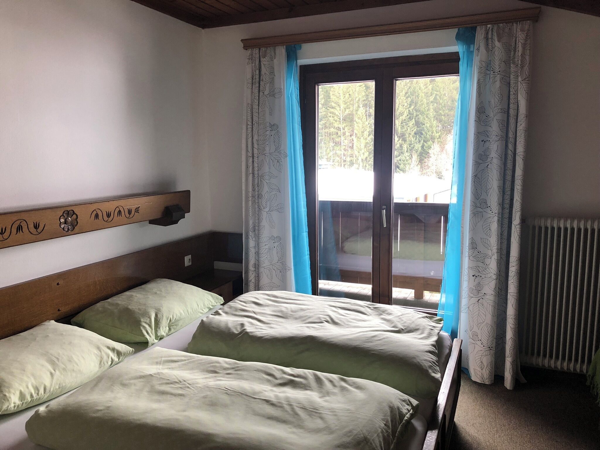 A bedroom with a wooden headboard, two beds with green bedding, a window with blue curtains, and a view of a deck outside.
