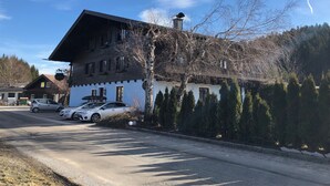 En tvåvåningsbyggnad med ett sluttande tak, parkerade bilar och en rad med vintergröna träd framför.
