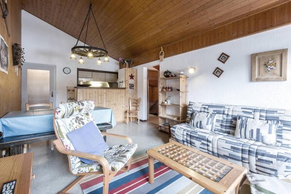 A cozy living room with a wooden ceiling, a blue sofa, a patterned armchair, a wooden coffee table, and a kitchen in the background.