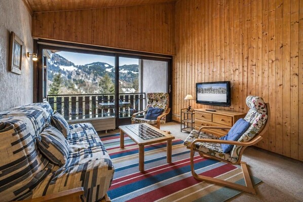 A cozy room with a sofa, chairs, a TV, and a large window offering a mountain view.