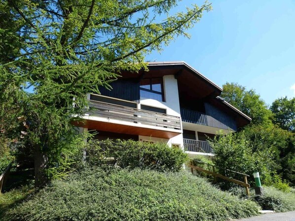 A modern two-story building with a balcony, surrounded by dense greenery.