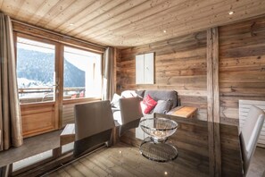 Interior de una cabaña de madera con sofá, sillas, una mesa y vistas a montañas nevadas.