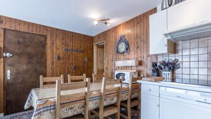 A dining area with wooden walls, a wooden table with chairs, a clock, and a kitchen area with white cabinets and a tiled backsplash.