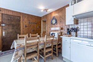 Ein Essbereich mit Holzwänden, einem Holztisch mit Stühlen, einer Uhr und einer Küche mit weißen Schränken und einer gefliesten Spritzschutzleiste.
