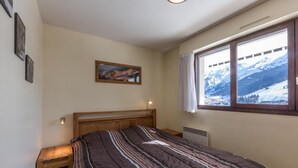 A bedroom with a wooden bed frame, a striped bedspread, a nightstand, a bedside lamp, a wall-mounted picture, and a window with a snowy mountain view.
