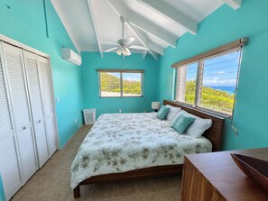 Une chambre à coucher avec un lit, un ventilateur de plafond, une fenêtre donnant sur un beau paysage et un panier à linge blanc.