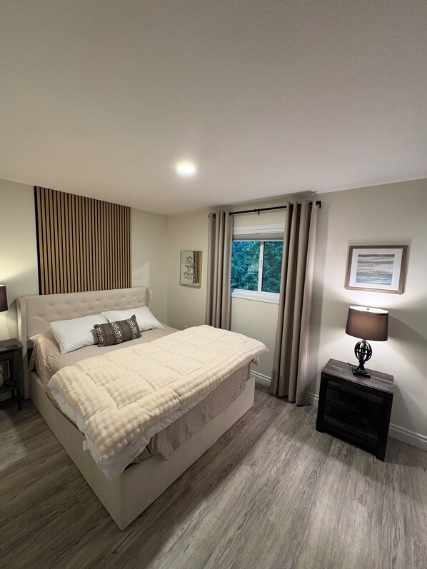 A bedroom with a large bed, a bedside table with a lamp, a window with curtains, and a wall-mounted artwork.