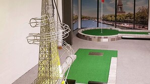 Ein Minigolfplatz mit einem grünen Putting-Bereich, einer metallenen Windmühlkonstruktion und einer Skyline mit einem markanten Turm im Hintergrund.