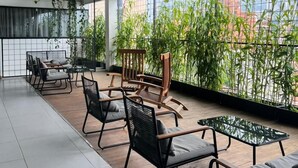 Un espacio moderno al aire libre con mobiliario de metal y madera, ventanas amplias y vistas a edificios y zonas verdes.