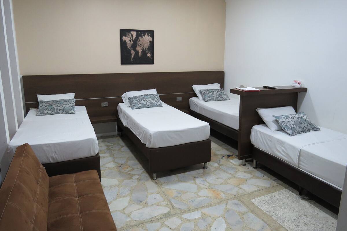 Une chambre d’hôtel avec trois lits simples, un canapé marron et une petite table sur laquelle est posé un panneau.