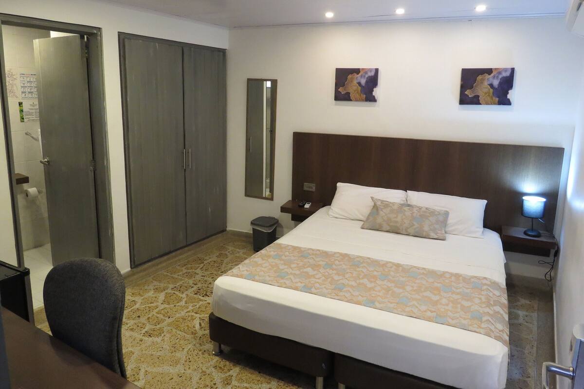 Une chambre d’hôtel avec un lit, une chaise, une armoire, un miroir et deux tableaux accrochés au mur.