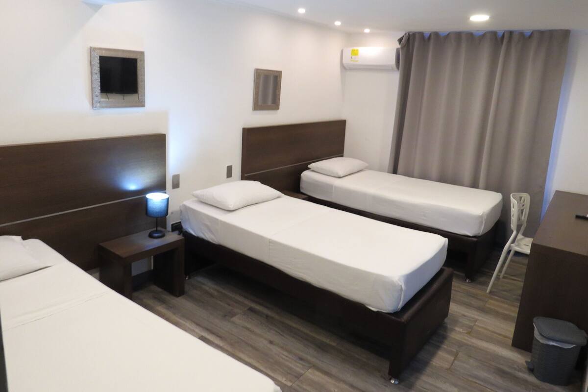 Une chambre d’hôtel avec deux lits simples, un bureau, une chaise, une table de chevet, une lampe, un appareil de climatisation et une petite fenêtre avec des rideaux.
