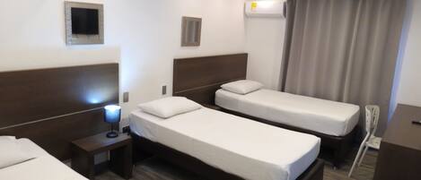 A hotel room with two single beds, a desk, a chair, a nightstand, a lamp, an air conditioning unit, and a small window with curtains.