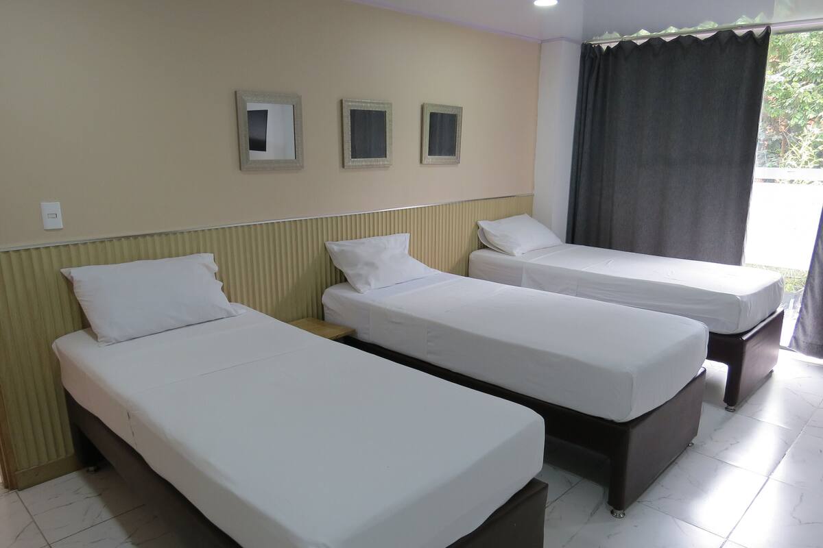 Une chambre d’hôtel avec trois lits simples, une fenêtre avec des rideaux et trois tableaux encadrés accrochés au mur.