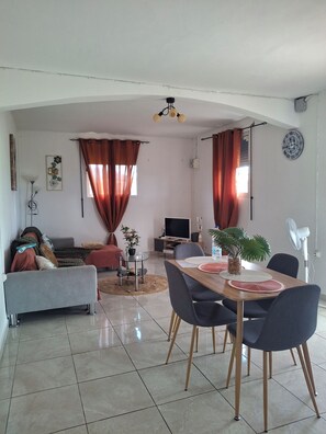 A living room with a sofa, dining table, chairs, a TV, and a fan. - Spacious country villa with parking and a large private garden. (Le Moule)