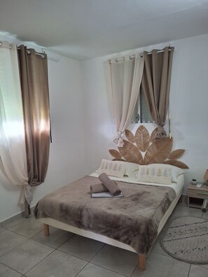 A bedroom with a bed, curtains, a headboard with a leaf pattern, and a bedside table with a lamp. - Spacious country villa with parking and a large private garden. (Le Moule)