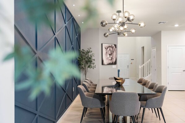 Un espace repas moderne avec une table en verre, des chaises grises, un lustre décoratif et un mur d’accent bleu.