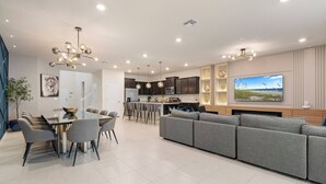A modern living room with a grey sofa, dining table with chairs, and a kitchen area with dark cabinets and a large TV. - 8BR house sleeps 16 with private pool (Davenport)