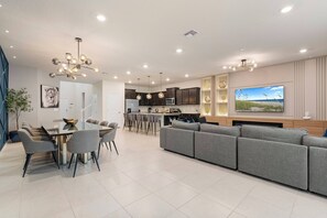 A modern living room with a grey sofa, dining table with chairs, and a kitchen area with dark cabinets and a large TV.
