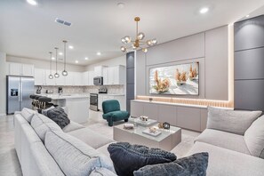 Una sala moderna con sofá, mesa de centro y un sillón color turquesa. Se ve, en el fondo, una cocina con armarios blancos y un comedor.
