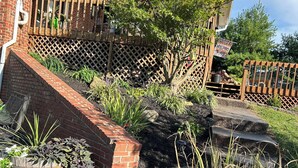 Een huis met een bakstenen gevel, een houten terras en een tuin met diverse planten en een kleine boom.