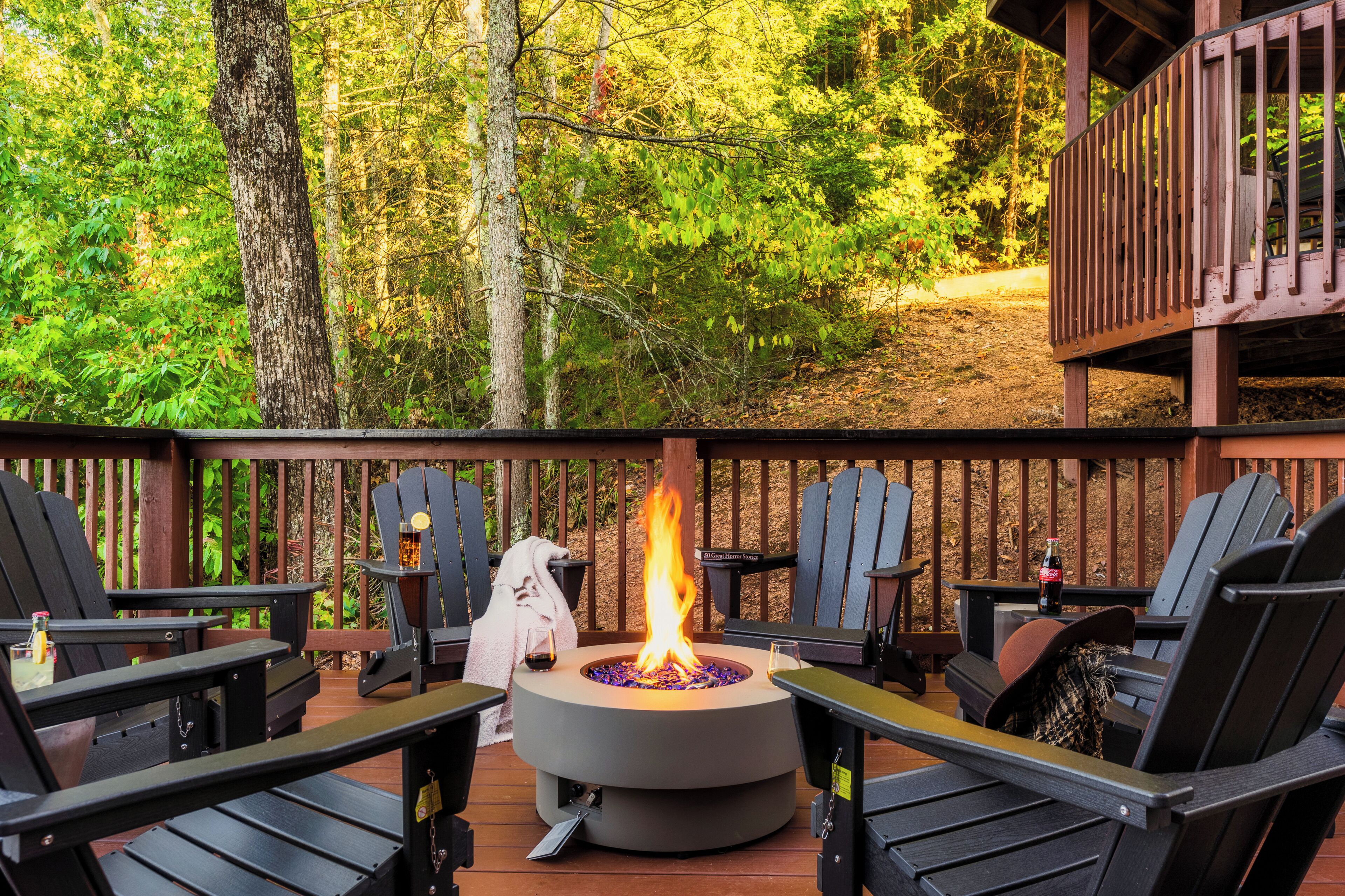 Una terraza con un brasero circular rodeado de sillas Adirondack negras, barandilla de madera y un bosque de fondo.