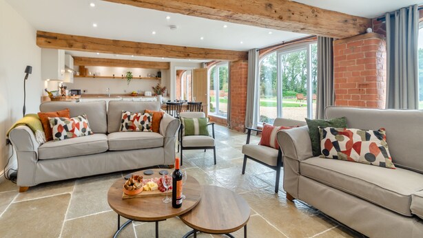 A spacious living area with a modern kitchen in the background, featuring wooden beams and large windows. - The Granary - UK46002 (Cranswick, near Driffield)