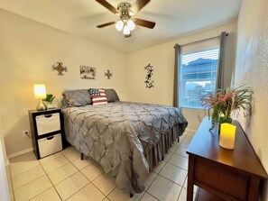 A bedroom with a bed, a nightstand, a lamp, a ceiling fan, a window with blinds, and a small table with a candle. - Tranquil Foothills Retreat  (Yuma)