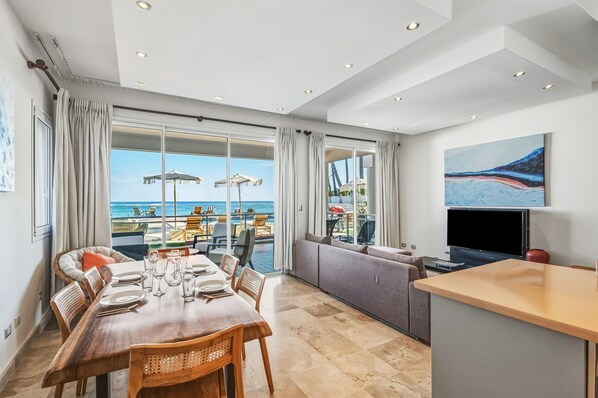 A modern hotel room with a dining area, a sofa, a TV, and a large window overlooking the beach. - Ocean Front Hot tub Pool Kite Beach Condo 101 (Kite Beach)