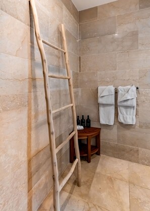 Un baño con baldosas de mármol color beige, una escalera de madera, una pequeña mesa de madera con artículos de tocador y dos toallas colgadas en la pared.
