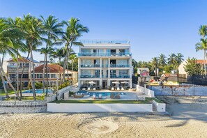 Un hotel de varios pisos con piscina, rodeado de palmeras y otros edificios.