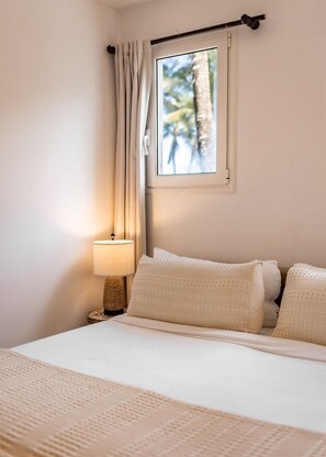 Un dormitorio con cama, ventana con vista a árboles, lámpara y mesita de noche.