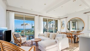 Amplia sala de estar con un ventanal que da a una piscina y una playa, amueblada con sofás blancos, sillas de mimbre y una mesa de comedor.