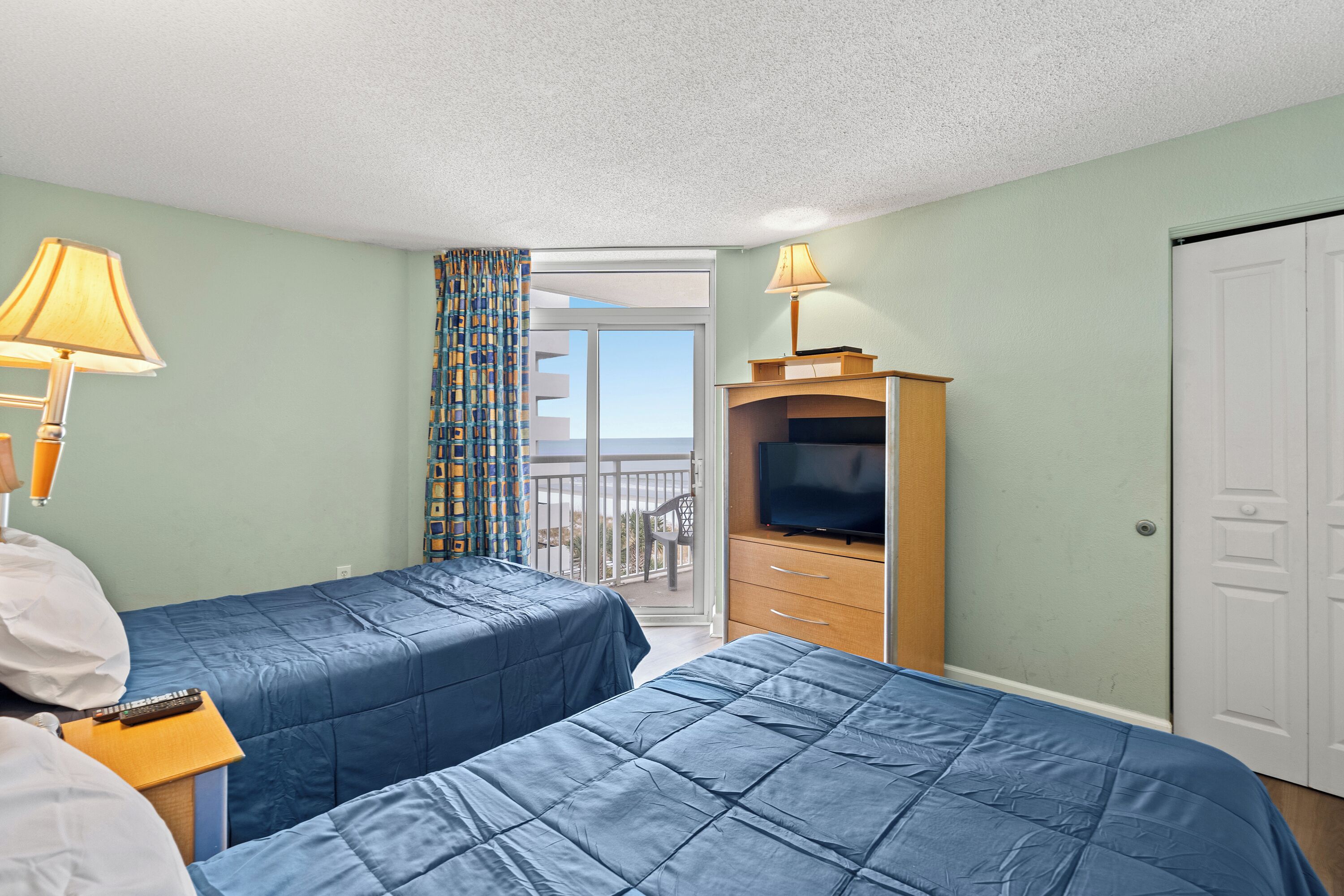 A hotel room with two beds, a TV, a desk with a lamp, and a balcony with a view.